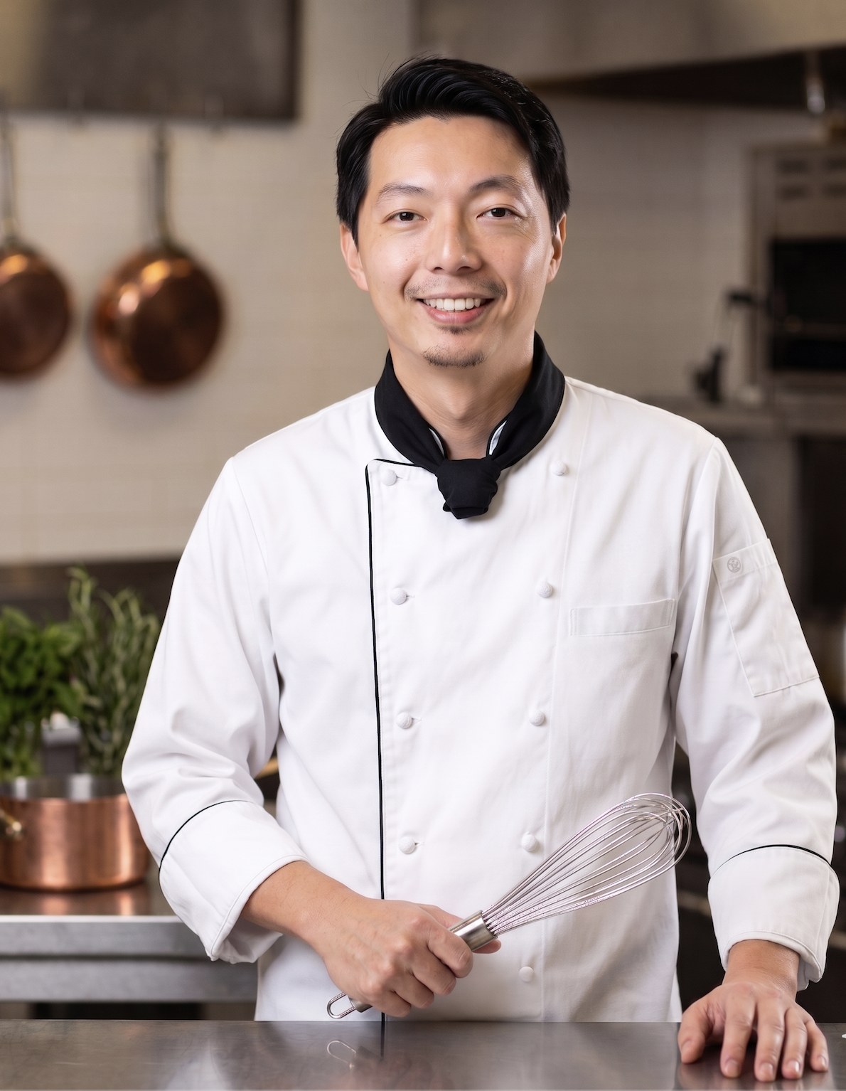 Chef Tsai smiling in his chef whites, holding a whisk in a professional kitchen